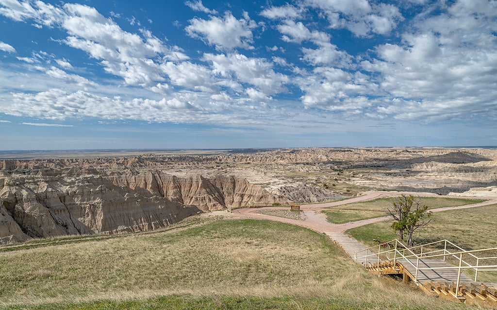 A blue sky with puffy white clouds over a basin of layered badlands formations and a grassy area with a fenced overlook looking out to the basin, Badlands National Park, South Dakota