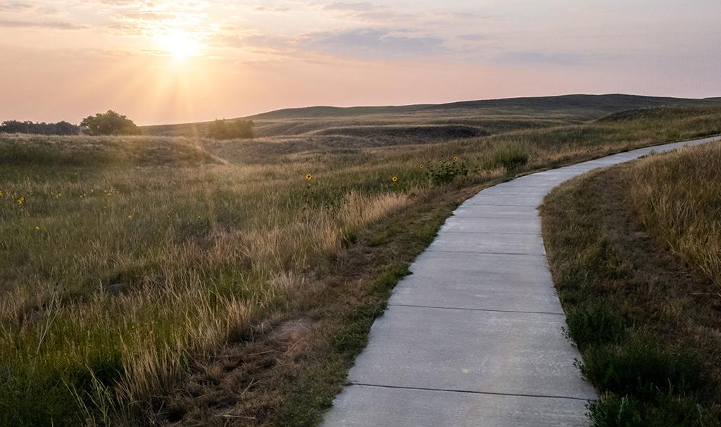 Sunrise sky over softly lit cement sidewalk style hiking trail. The trail slopes up and to the right around a hill. Several rolling grassy hills and a couple distant trees to right at Agate Fossil Beds National Monument.