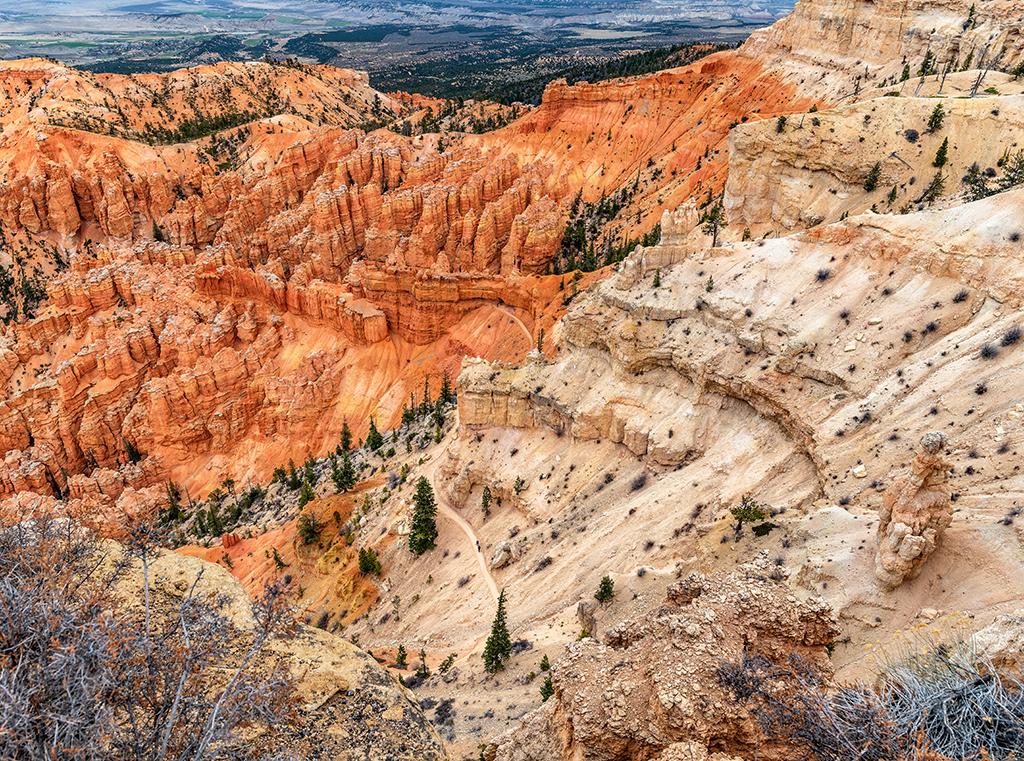 The tiny specks of two hikers on the trail surrounded by light beige and rust-red rock formations at Bryce Point, Bryce Canyon National Park