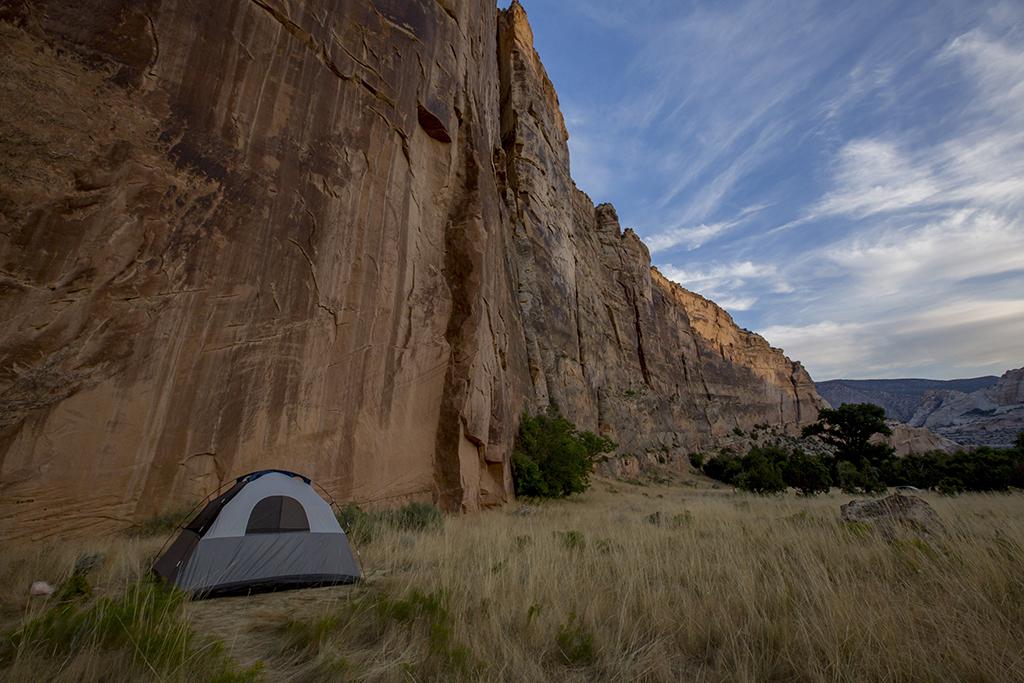 A tent set up against a high, red-rock cliff wall with a patch of blue sky and clouds overhead, Dinosaur National Monument