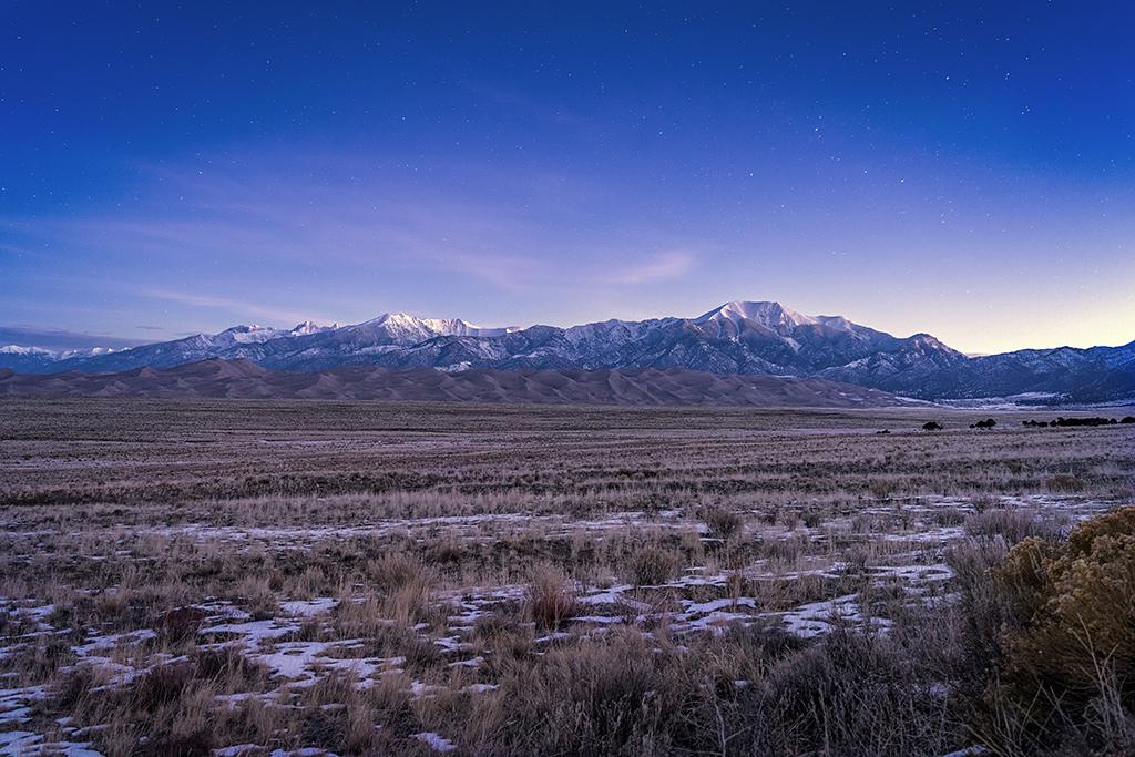 The winter night sky with a few remaining stars as the landscape begins to brighten with the coming sunrise over mountains and giant sand dunes at Great Sand Dunes National Park and Preserve.