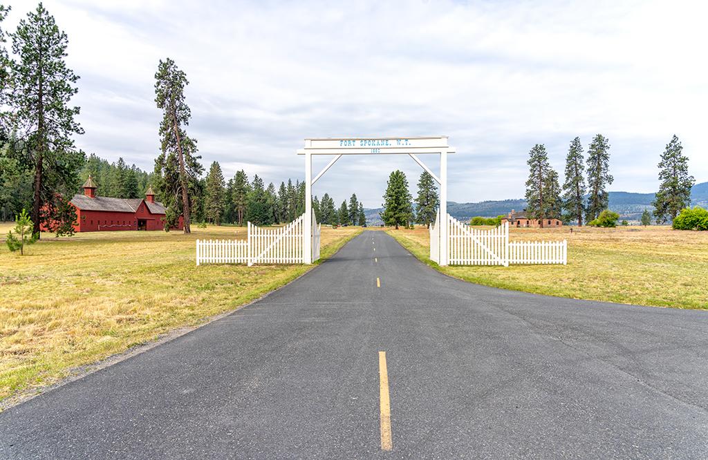 A road leading beyond a white entrance gate with blue lettering, and a field of grass on either side of the road. The red-painted wooden Quartermaster's Stable sits on the left side of the road beyond the gate at Fort Spokane in Lake Roosevelt National Recreation Area.