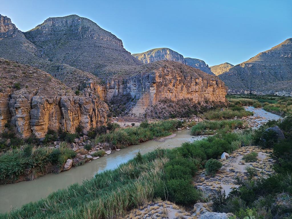 A view of the Rio Grande from a cliff above the river. From top to bottom, the image shows blue cloudless sky, steep beige cliffs dotted with green desert vegetation, a silty-green river interrupted by a rock garden rapid and in island, and bright green giant cane growing on both sides of the channel along the Rio Grande National Wild and Scenic River.