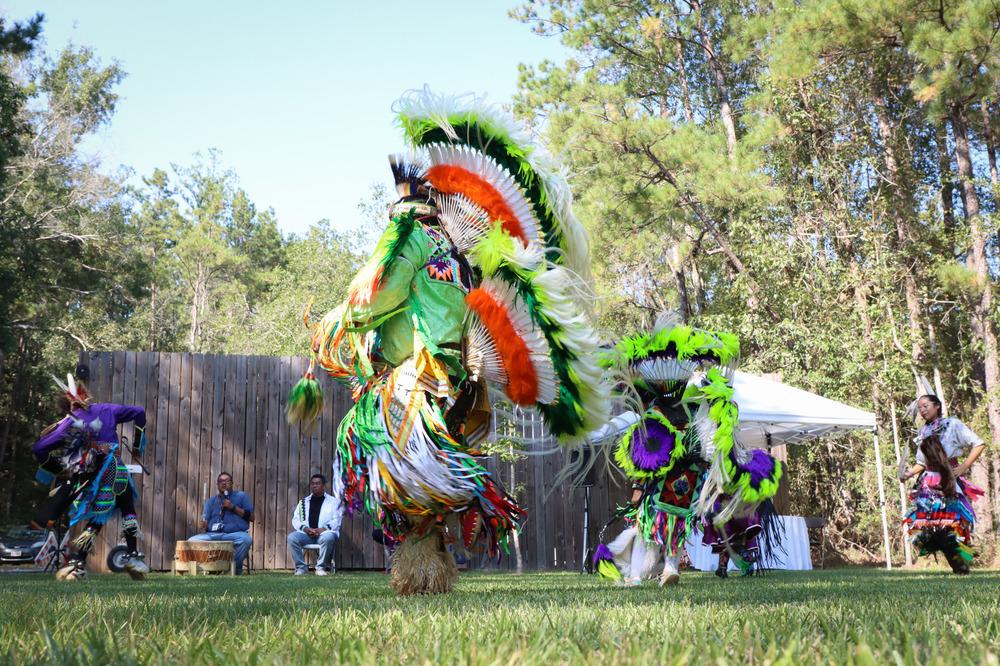 Alabama-Coushatta performers in brightly-colored regalia, displaying colors of green, orange, white, and purple, dancing on a lawn outside / NPS